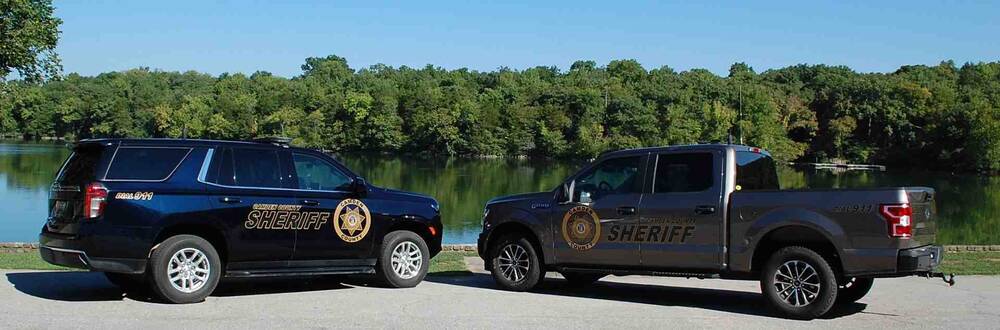Black sheriff suv and a gray sheriff truck parked next to a lake.