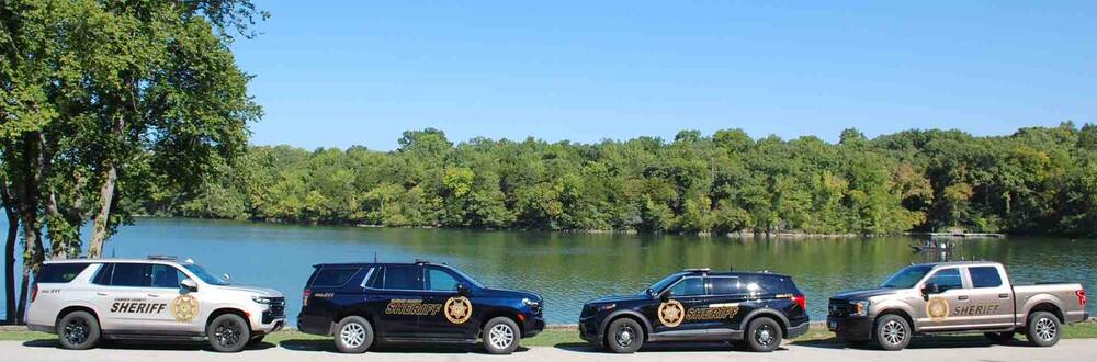 A line of four sheriff vehicles parked next to a lake.
